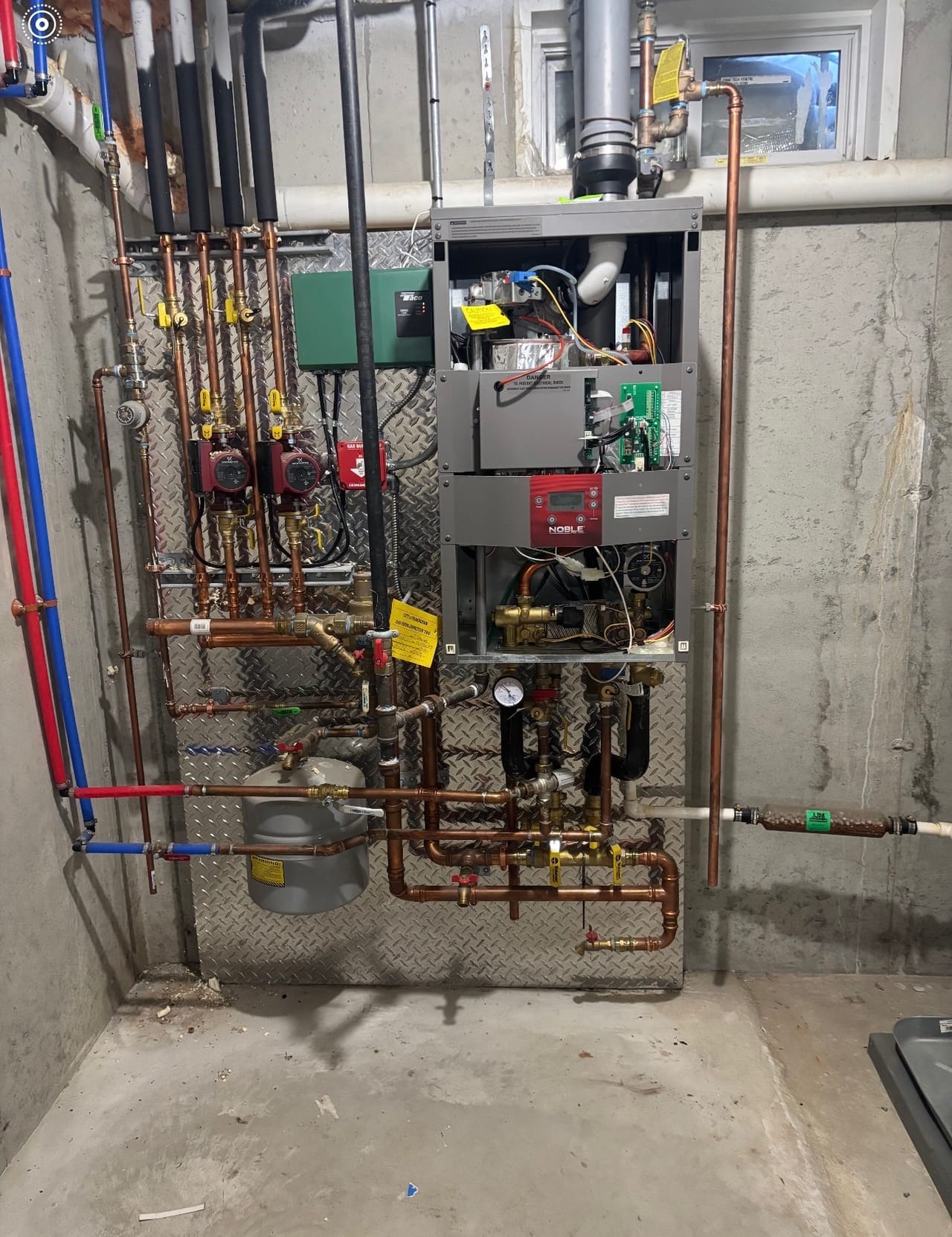 A Noble wall-hung gas boiler mounted on a diamond-plate backer board with twin circulator pumps and colour-coded supply lines