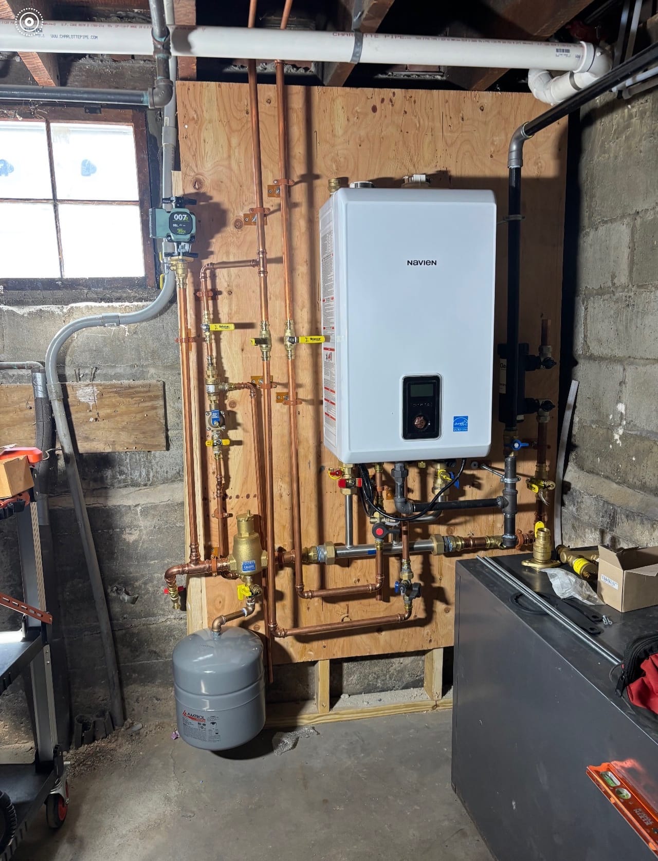 A wall-hung Navien tankless water heater installed on a plywood backer board with new copper supply lines and an expansion tank